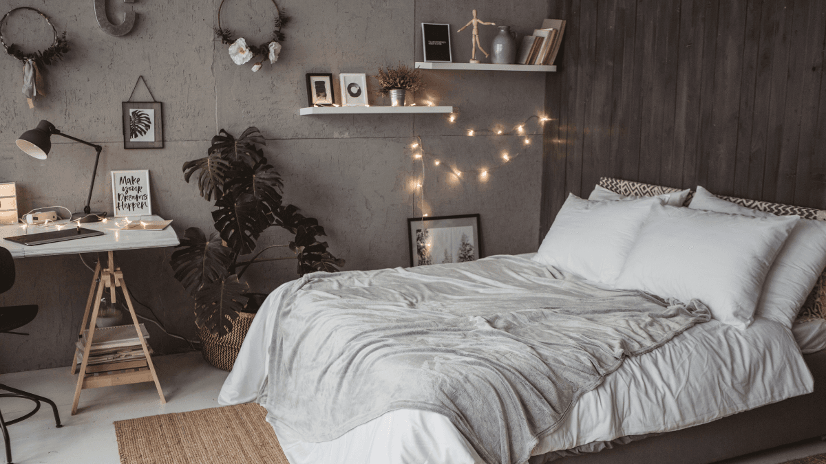 Modern bedroom with white bedding, gray throw blanket, indoor plants, wall shelves, string lights, and a compact workspace.